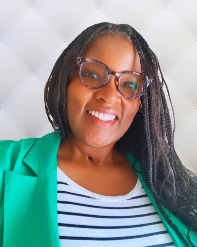 Aisha Adkins, a Black woman in her 30s, smiles at the camera. She is wearing a green blazer and a white top with black stripes. Her hair is in wavy braids.